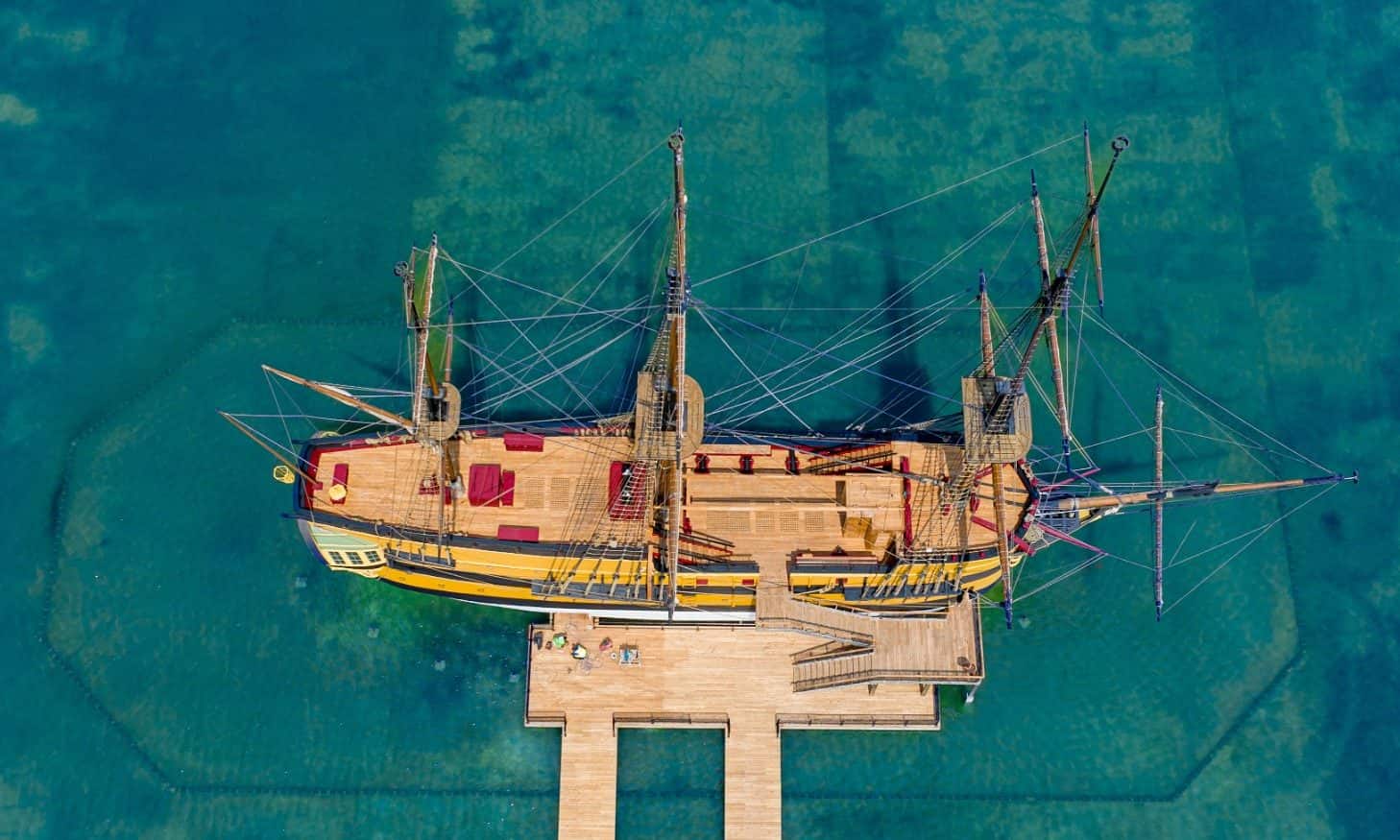 Aerial view of a three-masted ship moored at a wooden pier in shallow turquoise water