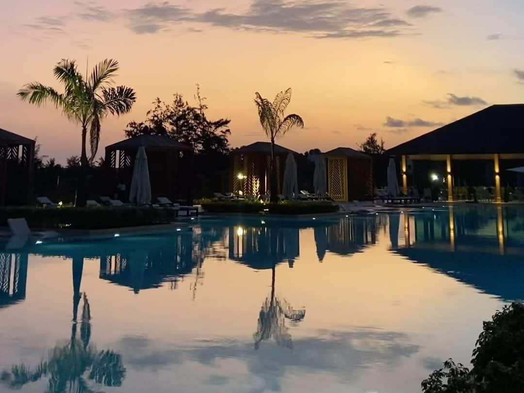 Pool of a Beninese hotel at dusk, palm trees and cabanas mirrored in the water